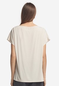 Kvinna med långt brunt hår som bär en beige kortärmad, löst sittande topp, sedd bakifrån mot en enkel vit bakgrund.