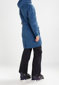Blaue, wasserdichte Jacke mit Kapuze und Gürtel, ausgestattet mit verstellbaren Manschetten und einem schlanken Design, kombiniert mit schwarzen Hosen und Schuhen.