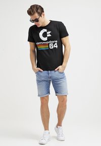 Camiseta de algodón negra con un logo blanco de "Commodore 64" y rayas de colores del arcoíris, combinada con pantalones cortos de mezclilla azul claro y zapatillas blancas.