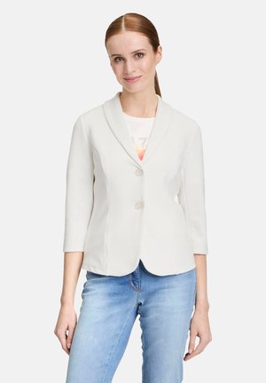 Witte, gestructureerde blazer met een sjawl kraag, driekwartmouwen en twee knopen; gedragen over een lichtgekleurde blouse en blauwe jeans.