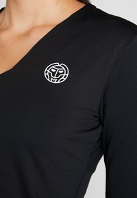 Svart topp med design av V-ringning, tillverkad av ett mjukt tyg. Har en vit cirkulär logotyp på vänster bröstområde.