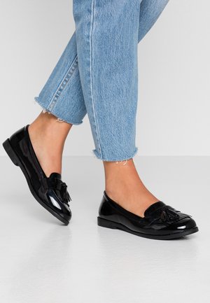 Voeten met glanzende zwarte loafers met kwastjes, gecombineerd met cropped lichtblauwe jeans met versleten zoom, op een effen witte vloer.