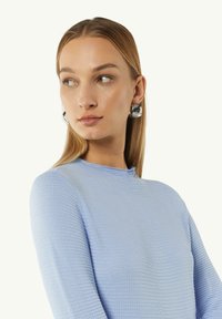 Hellblauer strukturierter Pullover mit hohem Kragen und langen Ärmeln. Das Model trägt große, auffällige Ohrringe mit einem kontrastierenden Design.