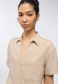 Camicetta beige a maniche corte con un tessuto testurizzato, chiusura frontale con bottoni e due tasche sul petto. Presenta un collo a punta.
