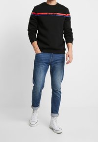 Černá mikina s červeným a modrým pruhem přes hrudník, kombinovaná s modrými džínami a bílými vysokými teniskami. Neformální střih a design.