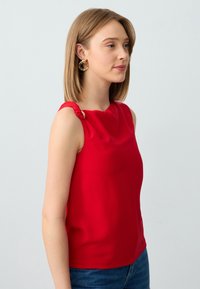 Top rosso senza maniche con scollatura drappeggiata e dettaglio alla spalla annodato. Tessuto liscio, design aderente, abbinato a jeans blu.