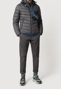 Man wearing a gray puffer jacket, dark gray pants, green and gray sneakers, and a blue crossbody bag standing with hands in pockets.