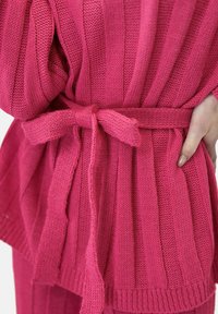 Różowy sweter z dzianiny o ryflowanej fakturze, związywany w talii paskiem i otwartym przodem. Materiał wydaje się być miękki i tkany.