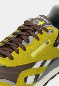 Αθλητικά παπούτσια Reebok με κίτρινη σουέτ επένδυση, καφέ συνθετικά στοιχεία, γκρίζες επενδύσεις και καφέ κορδόνια με λεπτομέρειες λογότυπου στη γλώσσα.