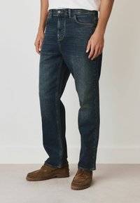 Mörkblå denimjeans med avslappnad passform, med dragkedja, framfickor och subtila blekningar. Matchas med bruna mockamokasin-skor.