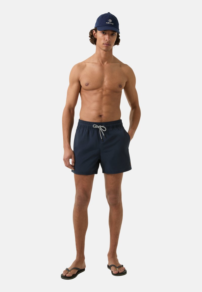 Marineblaue Badehosen mit einem elastischen Bund und Kordelzug, aus leichtem Material; getragen mit einer passenden Kappe und Flip-Flops.