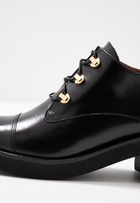 Černá kožená bota s lesklým povrchem, se zaoblenou špičkou a dekorativní zlatou hardware na tkaničkách. Zvýšená černá podrážka.