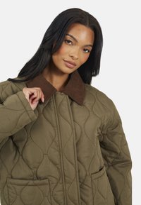 Veste matelassée de couleur vert olive avec un col en velours côtelé marron, dotée de poches latérales et d'un motif de vague texturé.