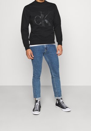 Sweat-shirt noir avec un logo Calvin Klein en relief, associé à un jean skinny bleu et des baskets montantes noires avec des accents blancs.