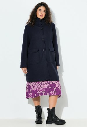 Abrigo de mezcla de lana azul marino con un alto cuello, cierre de botones y dos bolsillos laterales. Combinado con un vestido estampado en púrpura y rosa y botas negras.