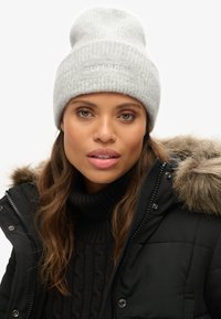 Gorro de punto acanalado gris con vuelta en el dobladillo, que presenta un logotipo sutil. Se lleva con una chaqueta acolchada negra y un suéter negro texturizado.