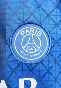 Blå atletisk trøye med diamantmønster, med et brodert sirkulært logo som viser "PARIS SAINT-GERMAIN" i hvitt.