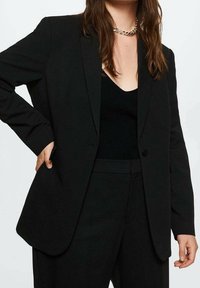 Black tailored blazer with a single button closure, notched lapels, and side pockets, made from a smooth fabric with a relaxed fit.