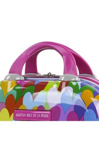 Maleta dura colorida con un acabado brillante, que presenta un patrón de corazones. Tiene asas rosas y herrajes grises, con un cierre de cremallera.