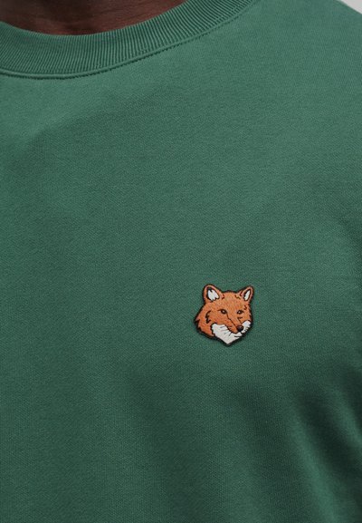 Πράσινο πουλόβερ με στρογγυλή λαιμόκοψη και ένα μικρό κεντημένο πορτοκαλί κεφάλι αλεπούς στην αριστερή πλευρά του στήθους, φορεμένο από ένα άτομο.