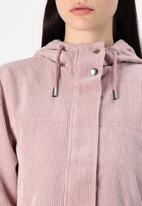 Lys rosa cordfløyelsjakke med hette, med glidelås, sølvfarge detaljer og ribbet tekstur.