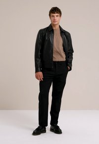 Giovane uomo che indossa una giacca di pelle nera, una camicia beige, pantaloni neri e scarpe nere, in piedi contro uno sfondo chiaro e semplice.