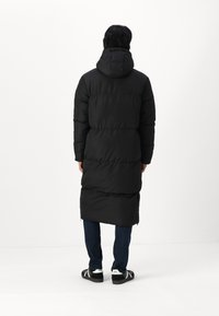 Zwarte lange pufferjas met een capuchon, voorzien van geborduurde stukken en een rits aan de onderkant, gecombineerd met blauwe spijkerbroeken en zwarte sandalen.