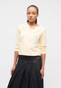 Νεαρή γυναίκα με πλεξούδες, που φοράει ένα εκρού πουλόβερ με κουμπιά και μπλε ανθισμένη κεντητή διακόσμηση, καθώς και μια ψηλόμεση σκούρα πλισέ φούστα.