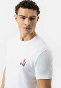Witte katoenen t-shirt met een kleine, kleurrijke afbeelding van een persoon in een rode outfit, die contrasteert met de stof.