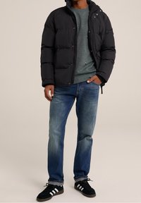 Man som bär en svart puffjacka, grå sweatshirt, blå jeans och svarta sneakers med vita ränder, står med en hand i fickan.