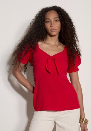 Kobieta o długich kręconych włosach, ubrana w czerwoną bluzkę z bufkami i wiązaniem z przodu oraz białe spodnie, stojąca z jedną ręką na biodrze.