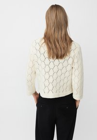 Donna con lunghi capelli castano chiaro che indossa un maglione bianco a maglia con motivo a foglia e pantaloni neri, mani in tasca, di spalle.