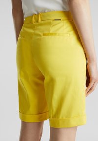 Pantalones cortos de algodón amarillos con dobladillos enrollados, que cuentan con dos bolsillos traseros y una textura suave. Las trabillas añaden detalle a la pretina.