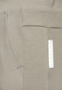 Graue Stoff-Jogginghosen mit elastischem Bund, seitlichen Taschen und einem weißen Etikett mit erhabenem Text. Das Material wirkt weich mit einer glatten Textur.