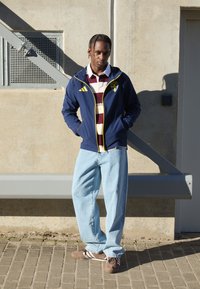 Jacket azul marinho com detalhes amarelos, camisa às riscas em bordeaux e branca, jeans azul claro e sapatilhas castanhas com riscas brancas.