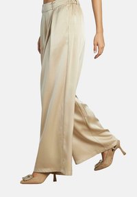 Pantalons en satin beige à jambes larges avec une taille haute et une ceinture élastique, associés à des talons beige avec une boucle décorative.