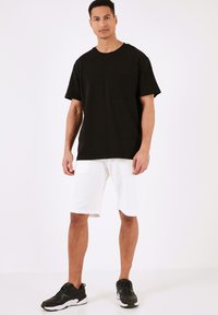Sort bomulds t-shirt med korte ærmer og en brystlomme, parret med hvide shorts til knæene og sorte sneakers. Glat tekstur.