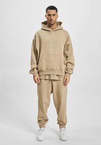 Sweatshirt beige à capuche avec une grande poche avant, superposé sur un pantalon de jogging beige ajusté. Complété par des baskets blanches, texture de tissu lisse.