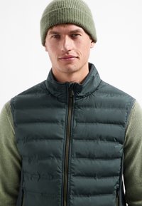 Donkergroen gevoerde vest met een rits aan de voorkant, hoge kraag en een gestructureerd beige shirt met lange mouwen eronder. Draagt een groene geribbelde beanie.