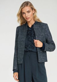 Kobieta ubrana w teksturowaną szaro-bluzkę, założoną na granatową bluzkę z falbanami oraz pasujące szerokie spodnie, na tle jasnego, jednolitego tła.