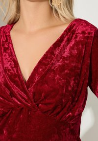 Czerwona aksamitna sukienka z głębokim dekoltem w literę V, marszczeniami w talii oraz miękkim, strukturalnym wykończeniem. Idealna na formalne okazje.