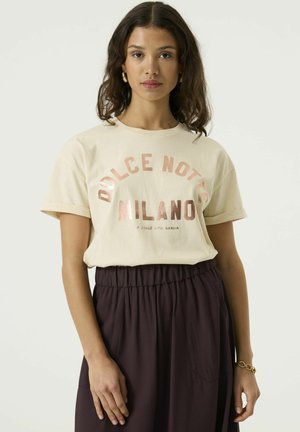 Mujer con cabello rizado oscuro que lleva una camiseta beige con el texto "Dolce Notte Milano" y pantalones oscuros de cintura alta, de pie contra un fondo liso.