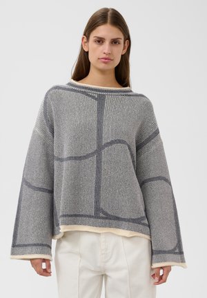 Jeune femme portant un pull en maille ample gris et blanc à motifs avec de larges manches et un pantalon blanc, debout devant un fond uni.