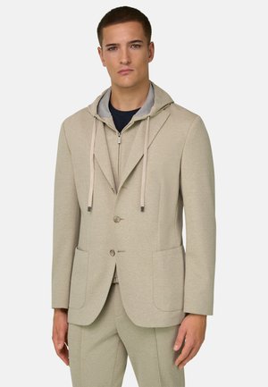 Jeune homme portant une veste de costume beige sur un sweat à capuche zippé gris clair avec cordons et un pantalon beige assorti sur fond blanc.
