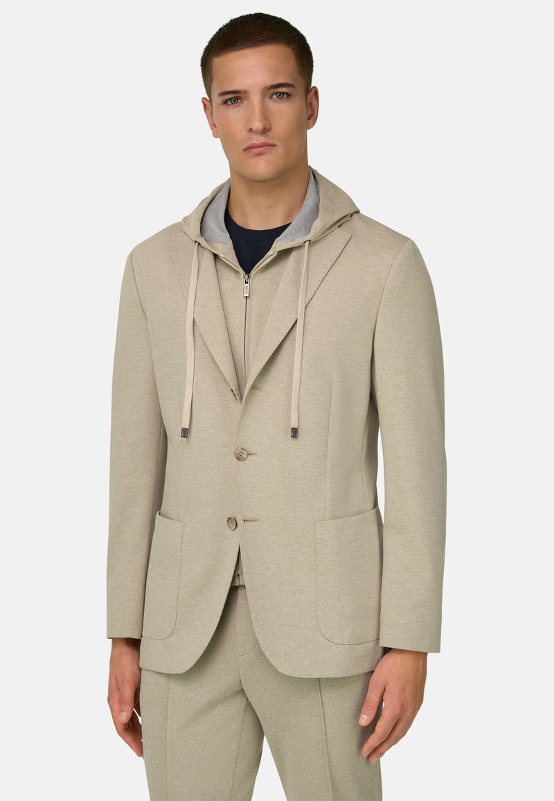 Junger Mann trägt eine beige Anzugjacke über einem hellgrauen Kapuzenpullover mit Reißverschluss und Kordelzug sowie passende beige Hosen vor weißem Hintergrund.