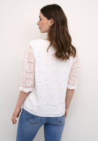Hvid blondebluse med trekvartærmer, der har et blomstermønster og en løs pasform. Sammen med blå jeans, der viser en afslappet stil.