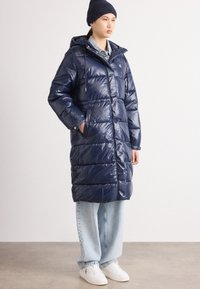 Geacă puffer de culoare bleumarin cu fermoar și nasturi tip capcană, cu glugă, mâneci lungi și buzunare laterale, purtată peste blugi decolorati și adidași albi.