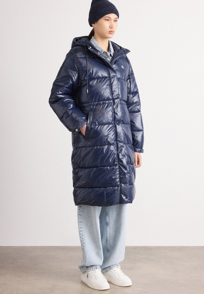 Geacă puffer de culoare bleumarin cu fermoar și nasturi tip capcană, cu glugă, mâneci lungi și buzunare laterale, purtată peste blugi decolorati și adidași albi.