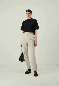 Vrouw die staat in beige cargojoggers, een zwart cropped t-shirt, zwarte sneakers en een kleine zwarte handtas tegen een effen achtergrond.