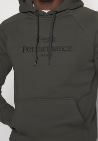 Felpa verde scuro con tasca frontale e cordini, ricamata con "Peak Performance Est. 1986" sul petto.